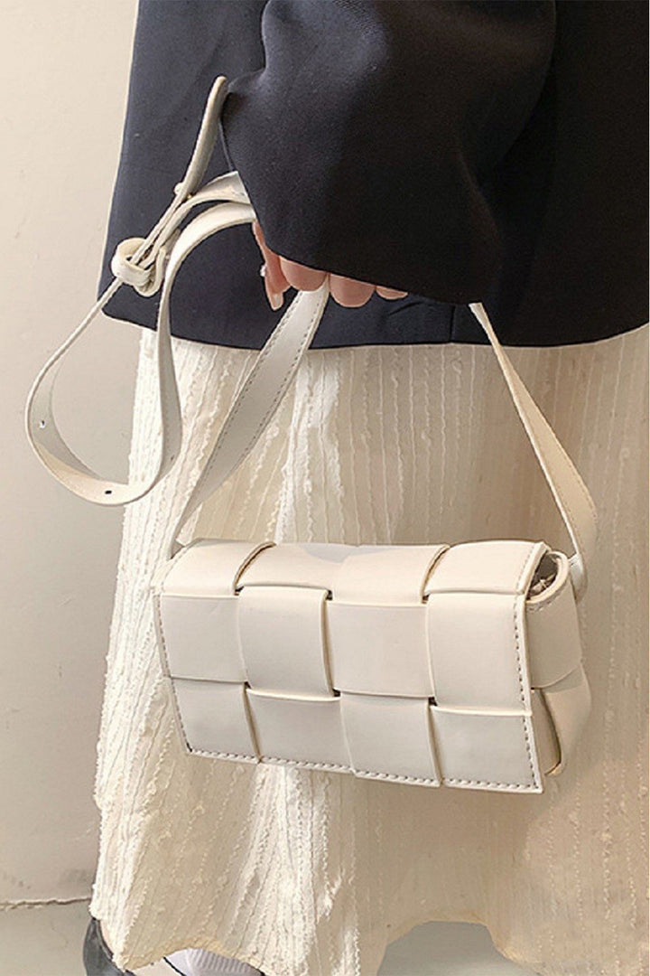 A person holds a Zenana Vegan Leather Woven Crossbody Bag with a long strap. Theyre dressed in a black jacket and textured, cream-colored skirt or dress against a neutral background.