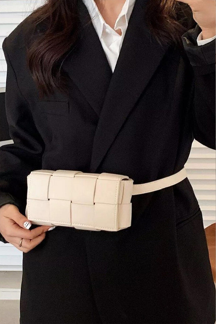 A person wearing a black coat sports the stylish Zenana Vegan Leather Woven Crossbody Bag, fastened around their waist. The eco-friendly white bag contrasts elegantly with the light-colored wall with horizontal lines behind them.