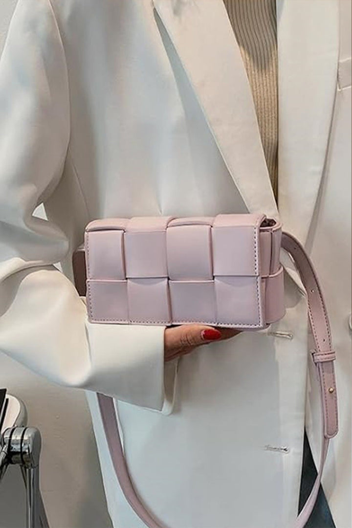 A person in a white blazer carries the Zenana Vegan Leather Woven Crossbody Bag in light pink. The eco-friendly accessory, with its shoulder strap, beautifully complements their red nail polish and beige top for a chic look.