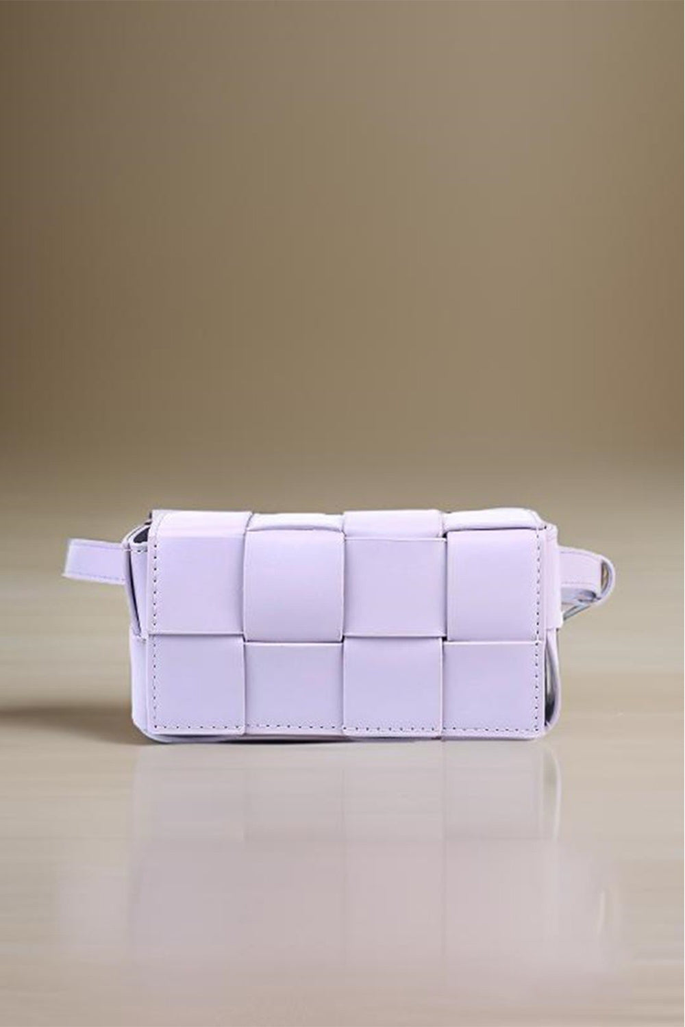 The Zenana Vegan Leather Woven Crossbody Bag, a stylish rectangular white clutch made from eco-friendly materials, is displayed on a reflective beige surface, highlighting its minimalist and modern design.