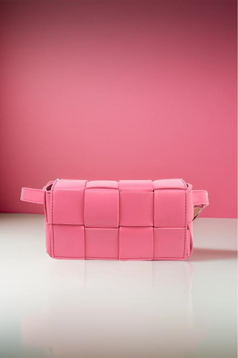 The Zenana Vegan Leather Woven Crossbody Bag, in pink, features a rectangular design with distinct square patterns and a side handle. Crafted from eco-friendly materials, it is displayed on a reflective white surface against a matching pink backdrop, blending style with sustainability.