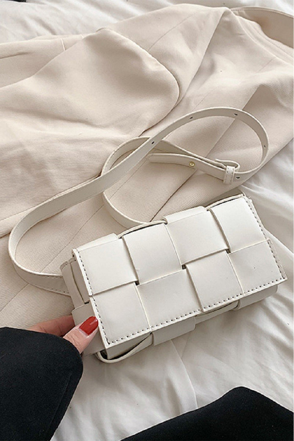 A hand with red nail polish holds the Zenana Vegan Leather Woven Crossbody Bag, eco-friendly and white, against a cream fabric. The bag has a long strap and rectangular shape.