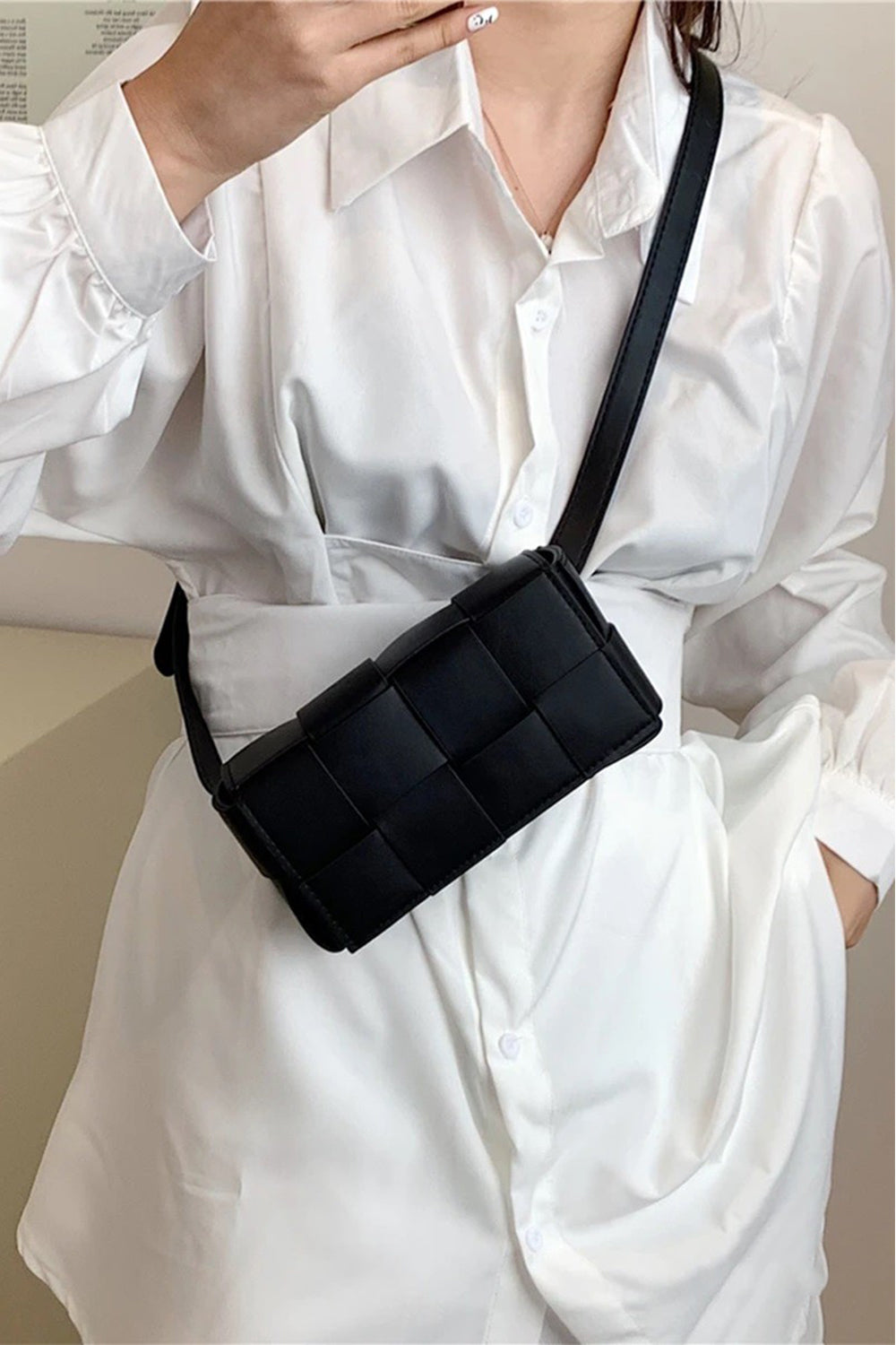 Wearing a white button-up shirt, a person holds a Zenana Vegan Leather Woven Crossbody Bag. This sleek, modern black bag features an eco-friendly grid-like pattern, with the hand resting near its top edge.