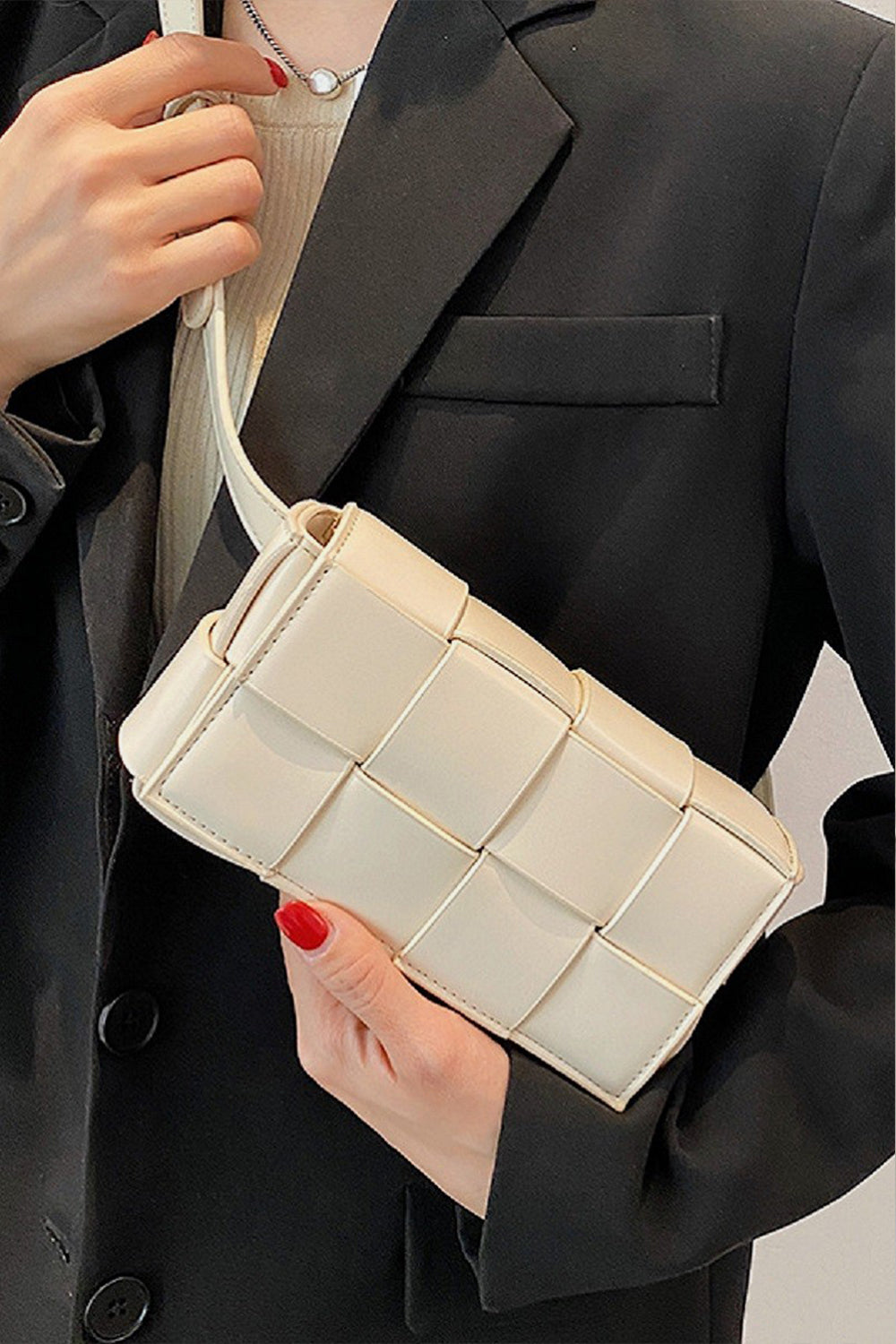 A person in a black blazer holds a Zenana Vegan Leather Woven Crossbody Bag, exuding style with red-painted nails and a round pendant necklace for an eco-friendly elegance.