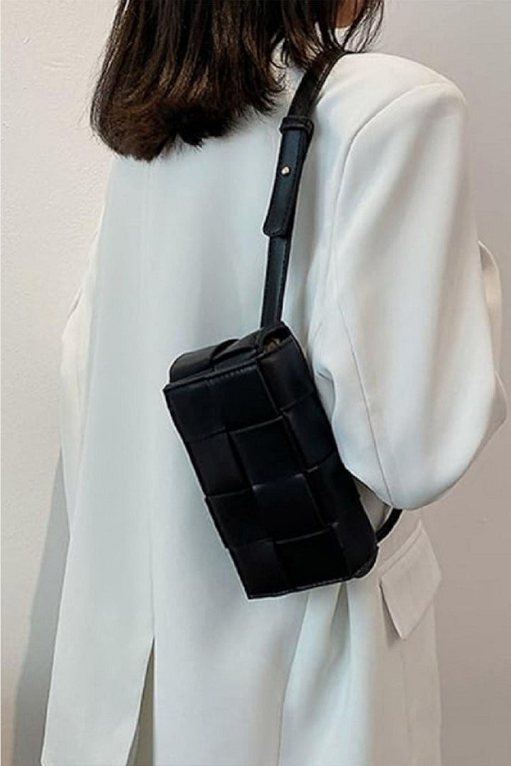 A person in a white blazer carries the Zenana Vegan Leather Woven Crossbody Bag. This eco-friendly bag, featuring a black textured checkered pattern, is worn over their shoulder against a plain, light backdrop.