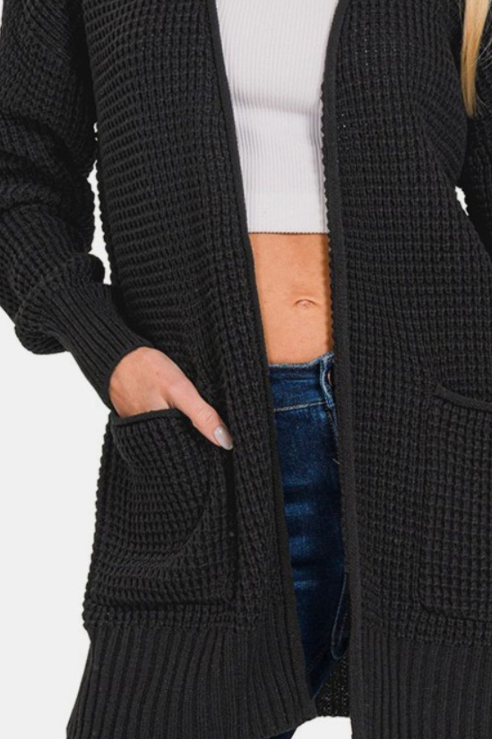 A person is wearing the Zenana Waffle Open Front Sweater Cardigan, a long black cardigan featuring front pockets over a white crop top and blue jeans. The cardigan's textured waffle knit enhances the stylishness of the outfit. With one hand in a pocket, this versatile wardrobe staple sets the perfect casual tone.