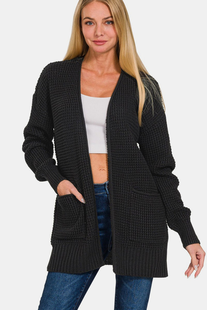 A person with long blonde hair is wearing the Zenana Waffle Open Front Sweater Cardigan in black over a white crop top and blue jeans. This versatile piece adds a textured waffle knit and front pockets to their outfit as they stand against a plain white background.