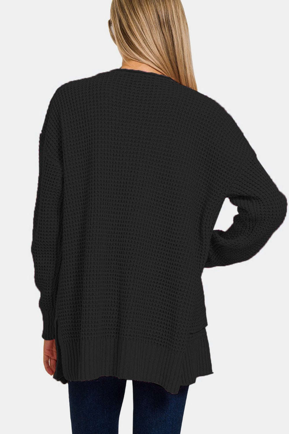 A person with long blonde hair, dressed in a Zenana Waffle Open Front Sweater Cardigan in black and dark blue jeans, is standing with their back to the camera. This versatile cardigan boasts a loose fit that extends below the waist. The background is plain white.