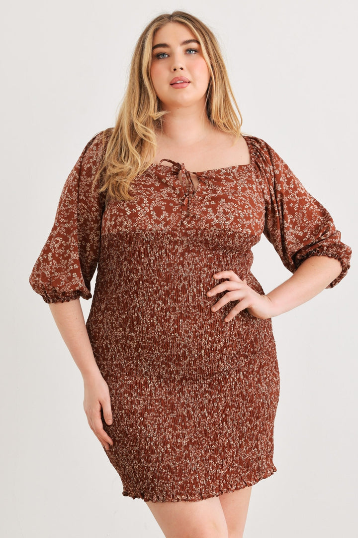 A person confidently poses with one hand on their hip, wearing the Zenobia Plus Size Smocked Printed Square Neck Mini Dress. This brown floral-patterned mini dress showcases puffed sleeves and a square neckline with a tie detail, set against a plain, light-colored background that accentuates its vibrant print.