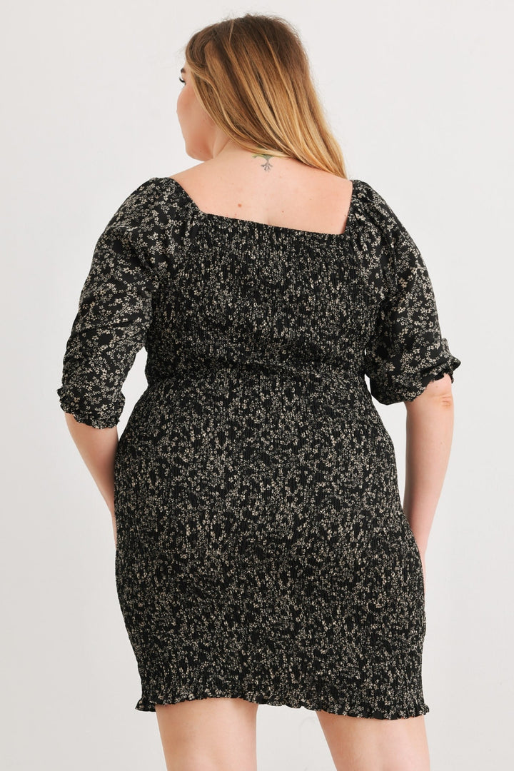 A person with long hair is donning the Zenobia Plus Size Smocked Printed Square Neck Mini Dress, characterized by its black and white floral design, square neckline, and puffed sleeves. They are facing away from the camera against a plain white background.