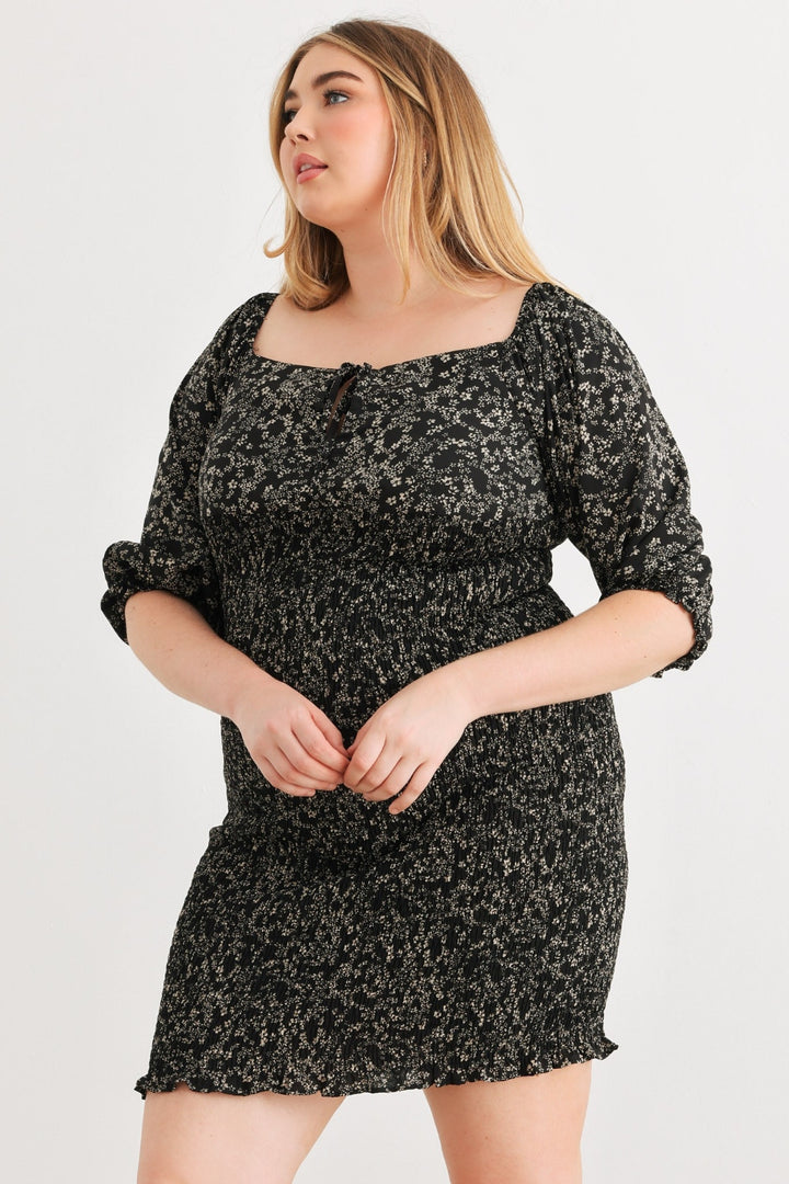 A woman stands against a plain white background wearing the Zenobia Plus Size Smocked Printed Square Neck Mini Dress. The dress, featuring a vibrant floral print on black fabric, has three-quarter sleeves and a smocked design. She gazes to the side with a neutral expression.